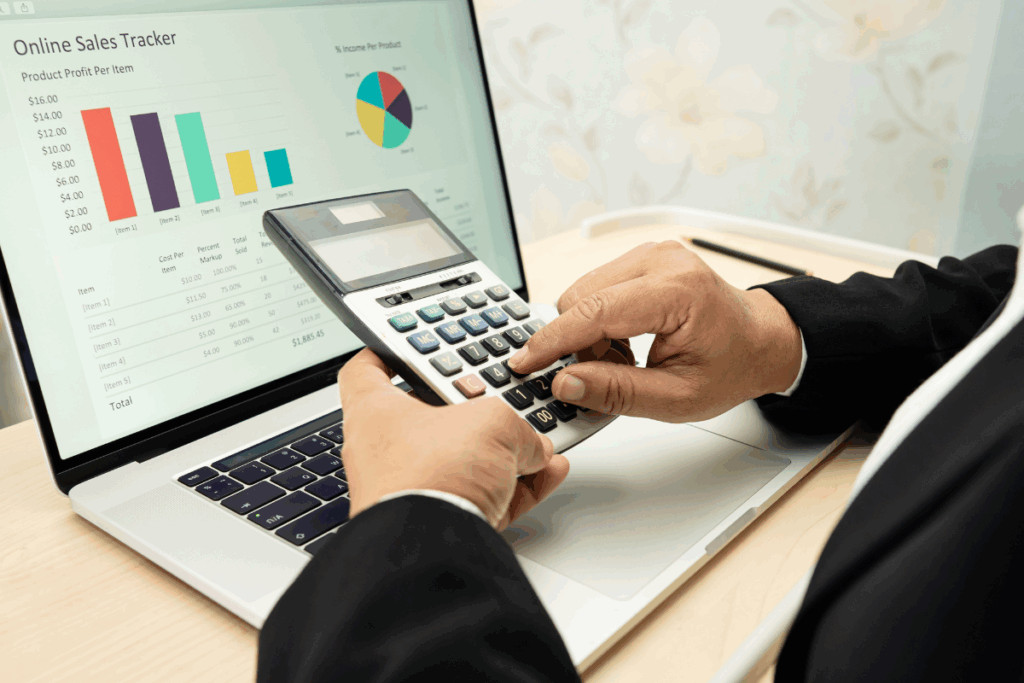 Close-up of a professional using a calculator in front of a laptop displaying sales and financial data, illustrating outsourced bookkeeping and financial reporting processes.