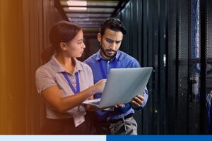 Two IT professionals, a man and a woman, working together in a server room while reviewing data on a laptop.