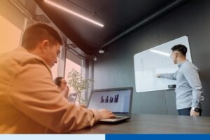 man in professional attire presenting using a whiteboard infront of audience with laptops in office