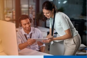 woman in office setting wearing corporate attire holding a tablet to man wearing headset with a desk computer