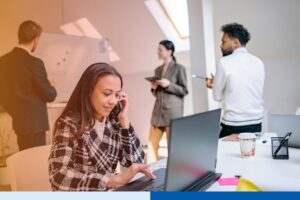 woman in office setting wearing corporate attire smiling while holding phone and working on a computer with people in the background