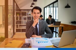 asian man in corporate suit attire smiling with paperwork and laptop in office