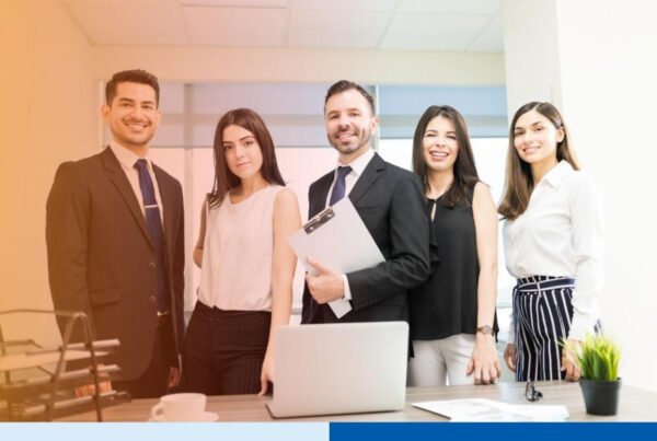 A diverse group of smiling business professionals standing together in a modern office behind a desk with a laptop, representing high-performing ⁠offshore teams and ⁠outsourced back-office staffing solutions.