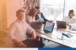 asian man in a corporate suit attire smiling with paperwork and laptop in office full of people