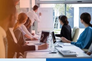man writing in whiteboard with people in corporate attire holding paperwork with laptops in office boardroom