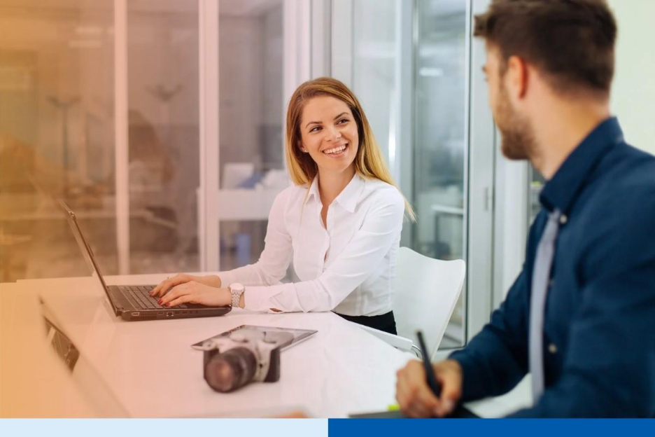 Two professionals collaborating in a modern office, smiling while discussing work on laptops 