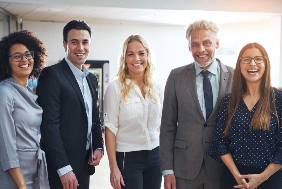 A diverse group of five smiling business professionals standing together in an office environment, dressed in business attire.