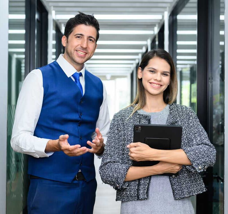 A smiling man and woman stand together in a brightly lit modern office hallway