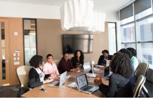A diverse group of businesswomen sitting around an office conference table with laptops, participating in a corporate meeting, representing customized BPO team management and global operations solutions.