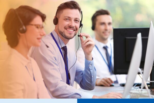 Three customer support professionals wearing headsets work at desktop computers, with one smiling toward the camera in a bright office.