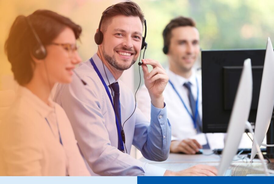 Three customer support professionals wearing headsets work at desktop computers, with one smiling toward the camera in a bright office.