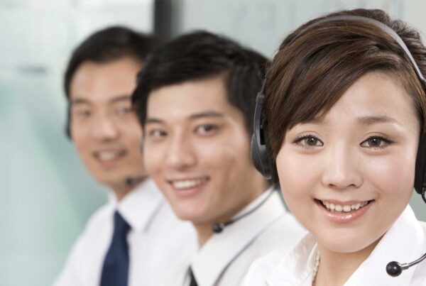 Three customer service representatives wearing headsets smile at the camera while seated together in a professional office setting.