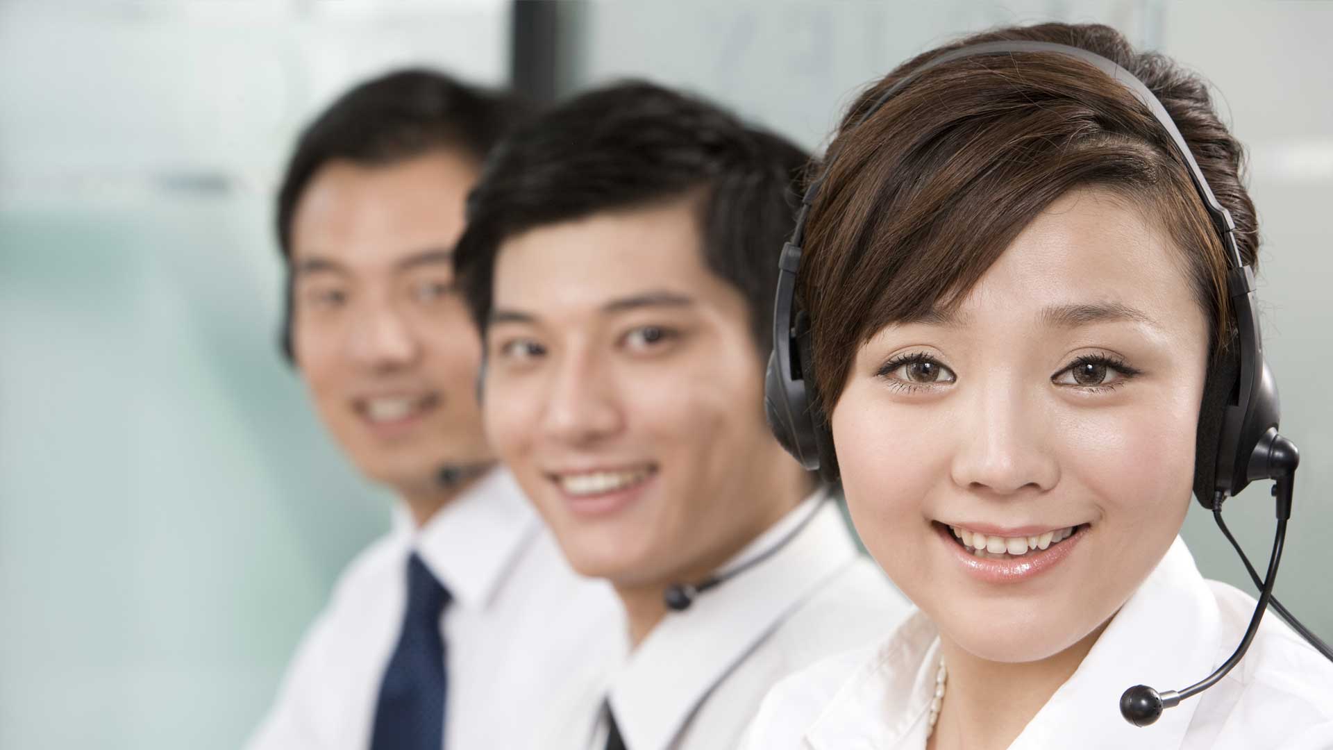 Three customer service representatives wearing headsets smile at the camera while seated together in a professional office setting.