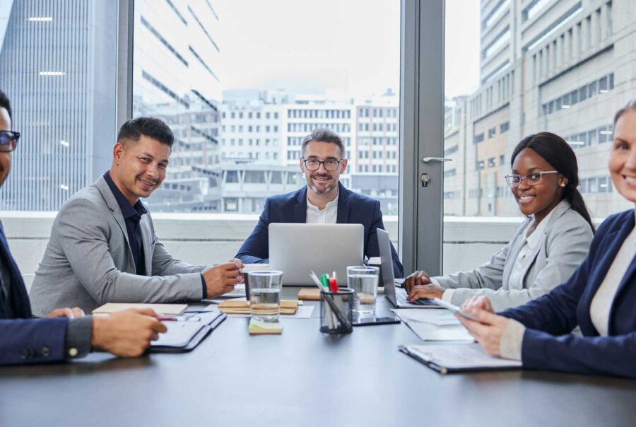 A diverse business team meeting around a conference table in a modern office with city views.