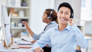 Smiling customer support professional wearing a headset at a computer workstation, with another headset-wearing colleague working in the background.