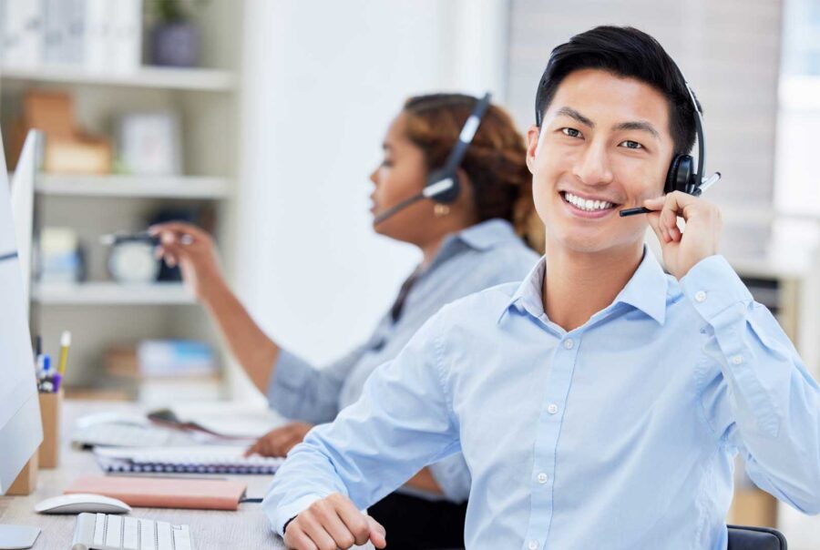 Smiling customer support professional wearing a headset at a computer workstation, with another headset-wearing colleague working in the background.