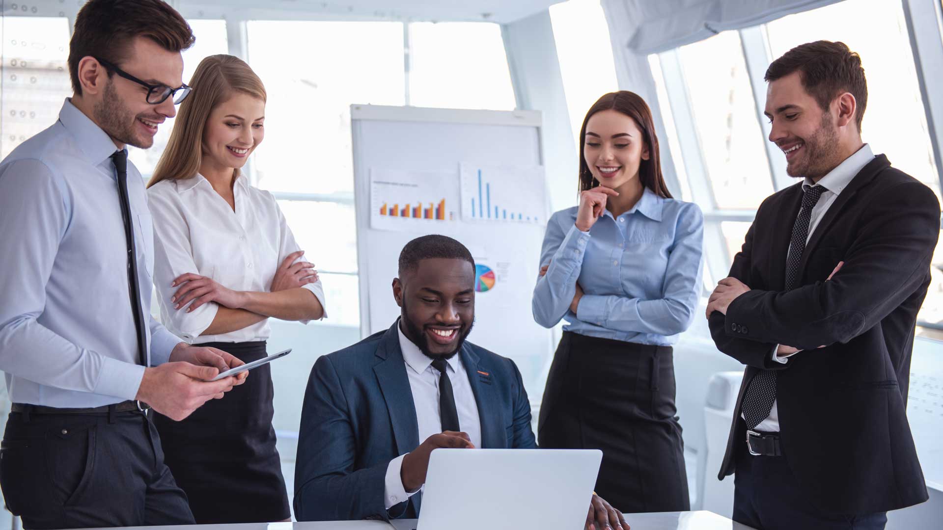 Five business professionals gathered around a laptop in a bright office.